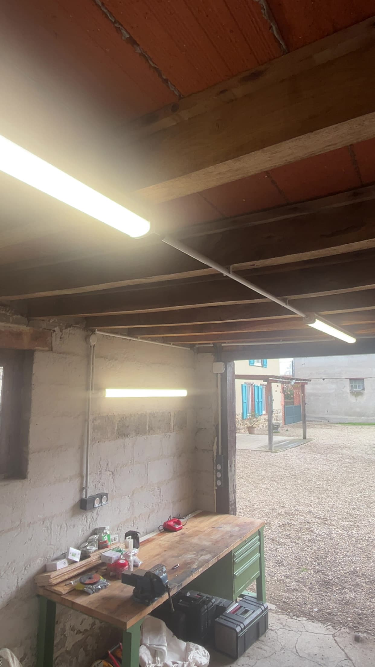 Atelier rustique avec établi en bois, éclairé par des néons, ouvert sur une cour extérieure.